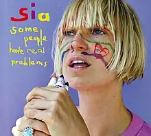 Sia stands against a light blue background, holding some markers in front of her face. There are drawings on her face. To the left of her, the name "Sia" and the title "Some People Have Real Problems" is stylized in a colorful, handwritten font.