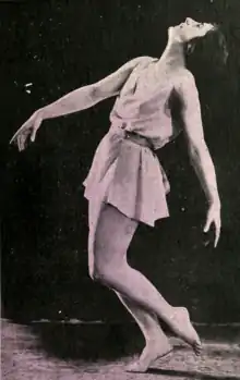 A white woman dancer wearing a short sleeveless dress, in a dance pose with both arms extended, both knees bent, and head thrown back
