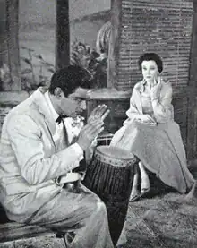 Indoor scene with young man playing a drum in the foreground, and seated youngish woman watching him; both are well-dressed