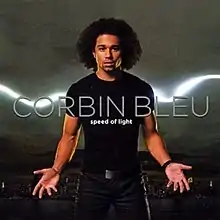 A man wearing a black t-shirt and jeans holding his hands out. Behind him is a stream of light and city skyline. The artist's name and album title are centered in front of the man, colored silver and white respectively.