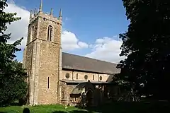 Stone church with a square tower. Unusually the windows in the nave are circular. The tower is on the left and dominates the picture. It has a crenellated flat top with stone pinnacles at each corner, narrow arched-top bell ports high up and very narrow slit windows lower down. The nave is receding left to right and is partly obscured by one of the two large yews that frame the picture. In shadow the porch can just be made out near the tower. The day is sunny and the sky mostly blue. The stonework of the church appears golden, similar to Cotswold stone. In the short grass of the foreground the top of a single gravestone can be seen.