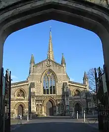 View through an archway of end of a church with a central door flanked by canopied niches containing statues. Arched window above the door and spire behind.