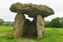 a grassy field in which three large upright stones support a stone slab roof