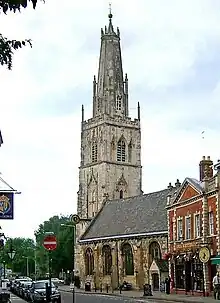 A stone church in a town seen from the southeast.  St the far end is a large tower surmounted by a truncated spire with pinnacles and, at the top, a ball finial