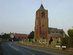 A curving road with buildings in the distance on the left and a church with a relatively large tower on the right. In front of the church are gravestones, a wall, and signs reading "Village Road" and "Long Lane"