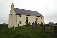 A large cream-coloured chapel, with two tall windows in the centre, a small belfrey and a slate roof. The sea is visible in the background.
