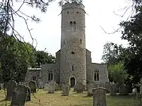 Front view of St Peter's church showing the late Anglo-Saxon flint round tower; Anglo-Saxon double bell opening; 14th century parapet, stringcourse and gargoyles. Set within a large churchyard with trees to both sides.