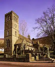 A stone church with a tall square tower, in the foreground is a stone wall and fountain