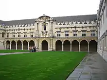  Facade of an ancient building, facing an expanse of grass. At ground floor level the building presents a long colonnade of arches, above which are rows of windows up to a crenellated roof line.