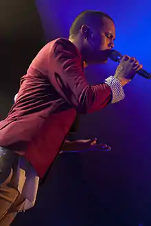 A man with brown skin wearing a red suit jacket and holding a microphone.