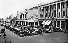 Black and white photograph of a business district with cars parked in the middle of the street and businesses lining the street