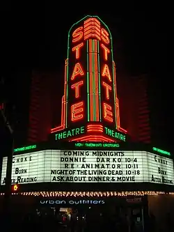 The neon marquee of a theater lists several notable cult films including Donnie Darko, Reanimator, and Night of the Living Dead.