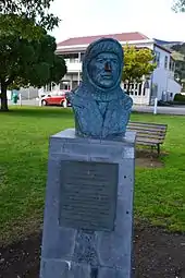 A bronze bust of a man wearing a balaclava, the bust on a plinth in a park area with a backdrop of a park bench and buildings in the far distance