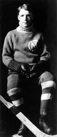 A young man in full hockey gear sitting.  He has short hair parted on his right side and is wearing a dark uniform with a light coloured "M" logo in a circle on his left breast