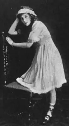 A young white woman leaning on a chair, wearing a short "babydoll" dress with short puff sleeves and a bonnet