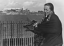 A photograph of a woman standing behind a fence, opposite of Alcatraz island. She is wearing a dark tassled shawl, which is blowing in the wind and her long hair is braided with beads, leather straps, and animal fur.