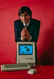 Steve Jobs resting his forearms on a Macintosh computer, in front of a red background.