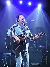 A man with dark hair and mustache playing guitar and singing into a microphone
