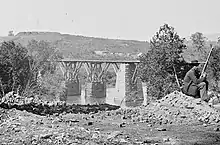 Black and white photo of a bridge, with a seated sentry at right and a fort crowning the height at upper left.