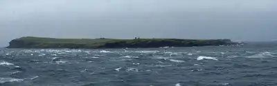 View of a low-lying island under grey skies, with rough water and cresting waves in the foreground