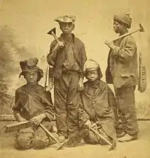 A studio portrait of four Afro-American climbing boys from New York, with brushes and scrapers, Two are standing and two are kneeling. They look between eleven and fourteen years old, wear rough clothes and battered hats and caps.