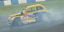A yellow and blue car spinning on a dirt track with a white man in overalls hanging from the side door looking to the rear.