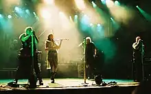 Four singers perform in the spotlights on stage. One is a female in shorts, a gauzy beige skirt and a leather belt, and plays the fiddle,  The males are wearing black sleeveless shirts, black pants and have military-looking gear. A rock band is in the background.