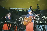 A man plays acoustic guitar in the centre of a stage, the black back wall of which is filled with the words "Spring & Airbrake" in white lettering. A female keyboardist is behind and to the left of the man, and in the background can be seen a male bassist and a drum kit.