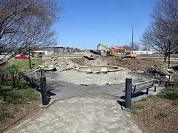 Construction equipment has begun to dig up concrete and level the earth beyond an area with a concrete fountain and wooden benches.