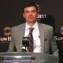 A color photograph of Neal Brown in a suit and tie at a podium.