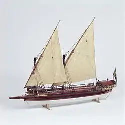 A side view of a model of a small galley with two masts rigged with lateen (triangular) sails. Its outrigger folded up and the oars stowed on the deck. The hull above the waterline is painted red with decorative details in gold and blue. The bow has a raised platform (rambade) armed with 3 small cannons.