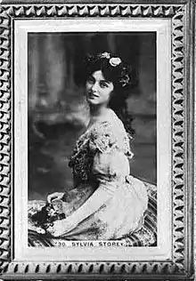 A young white woman, seated, wearing a gown. She has flowers in her dark hair. The image is framed and captioned "Sylvia Storey".