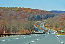 A divided highway in the middle of a late autumn wooded landscape curving up and down through a small valley with hills in the distance