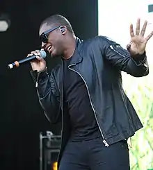 A black male wearing black sunglasses and dark jacket, shirt and trousers, sings into a microphone.