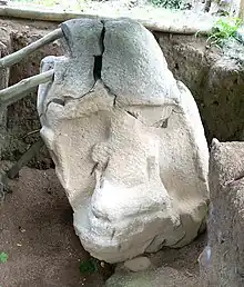 A stone sculpture sitting in an excavated trench. The monument is damaged with deep fracturing towards the top. It lists to the left and is braced with wooden poles. A niche is carved into the front of the monument and contains a badly eroded seated figure holding a featureless bundle in its arms.