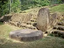 A squat standing stone with a flat front face set against a low stone-faced mound structure at right. The face of standing stone is inscribed but the details are not visible. In front of the stone, at left, lies a flat circular stone, set upon a flat grassy area. Dense vegetation is visible in the background.