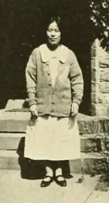 A young Japanese woman, standing outdoors in front of steps, wearing a light-colored dress and a cardigan