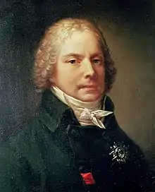 Portrait of a man, showing his head and shoulders. He is wearing a black jacket with a high-necked collar and a white shirt tied in a bow. His head is adorned by a curly blonde wig.