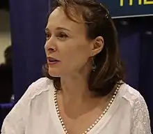 A bust-photo of a white woman with brown hair; she is wearing a white shirt and earrings, facing and looking to the camera's left.