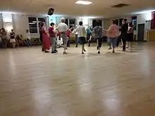 A small circle of dancers with one foot raised hold hands while several people look on.