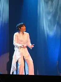 Turunen sings live onstage while seated on a stool. She is wearing a glittery white suit.