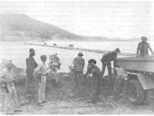 Six men in floppy hats shovel dirt. Two men wearing steel helmets and carrying pistols in holsters watch.