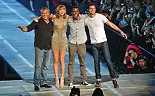 Four people—a middle aged white man in a striped black shirt and jeans, a young white woman in heels and a sparkling dress, a black man in a grey shirt and pants, and a white man in a white tee and jeans, posing together onstage