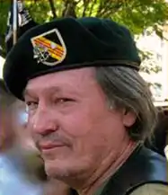 A white man with shoulder-length black hair tinged with gray, seen from his left, wearing a dark green beret with military insignia