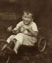 Toddler Ted Stevens rides on a tricycle. Stevens has a smile. The image is in sepiatone.