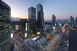 An evening rush hour shot of the intersection between the Teheran boulevard and Yeongdong highway from above. Multiple skyscrapers are visible, along with a pedestrian plaza and a stadium. In the background, apartment buildings and mountains are visible.