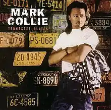 An image of Mark Collie wearing a white t-shirt and blue jeans, carrying a flannel shirt while next to a wall filled with Tennessee license plates. The artist's name and album title appear on the top-left of the cover, colored in white and orange respectively.
