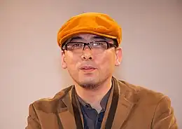 A bespectacled Tensai Okamura, in an orange corduroy cap