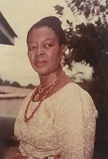 A Nigerian woman wearing a white blouse and red bead necklace. Standing outdoors.