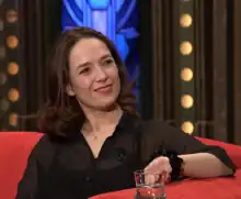 Tereza Kostková sitting on a red sofa, smiling and looking left of camera with a drink next to her hand